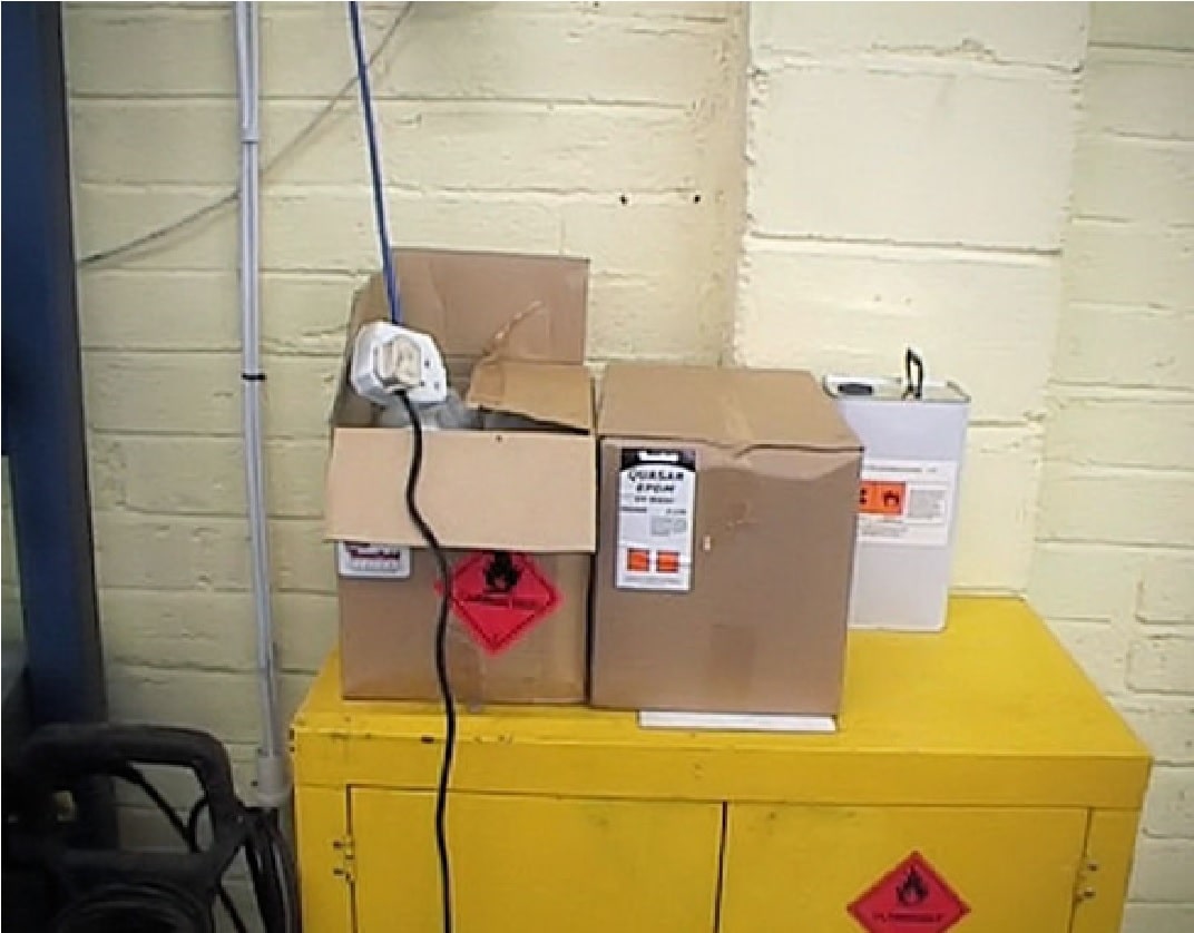 unsupported extension lead hanging over boxes of flammable substances which are stood on top of a yellow flammables storage cupboard